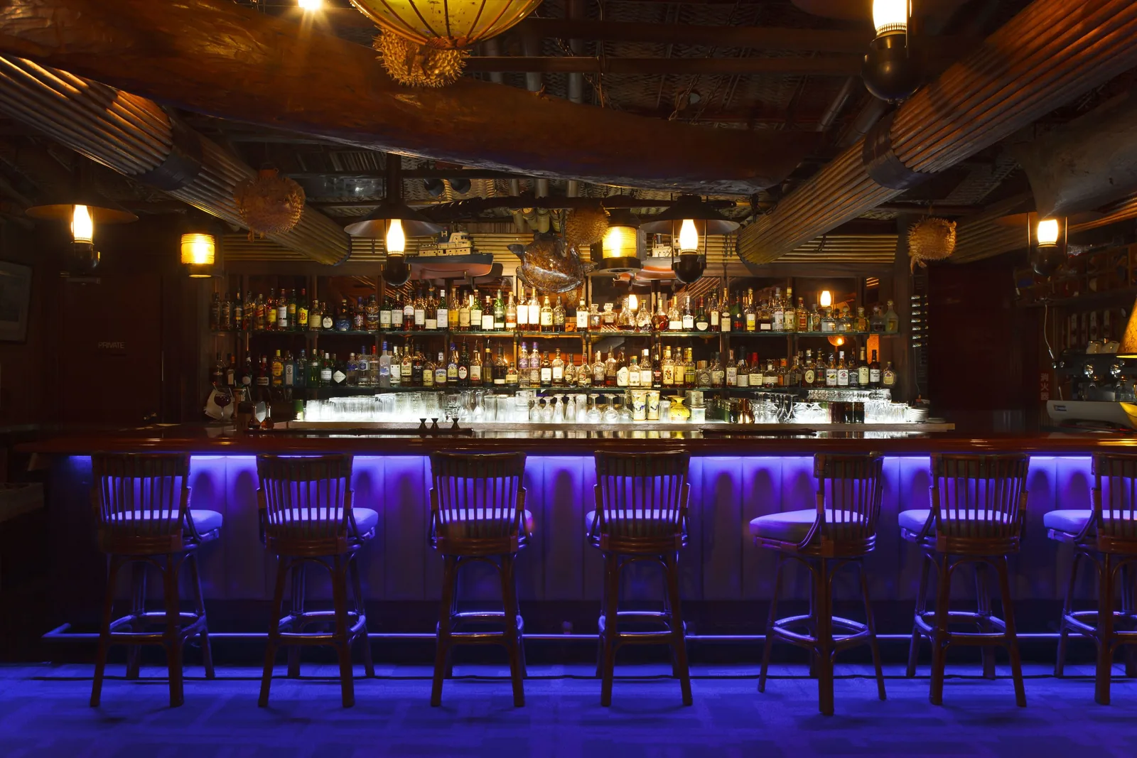 Hotel bar with a long wooden counter, high stools, shelves of bottles, warm pendant lights, and blue lighting below the counter