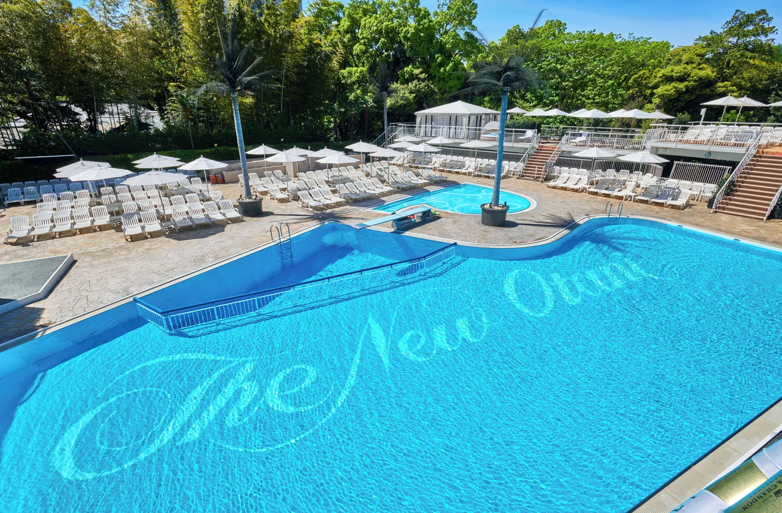 Large outdoor swimming pool and a smaller pool surrounded by rows of white loungers, umbrellas, palm trees, and greenery